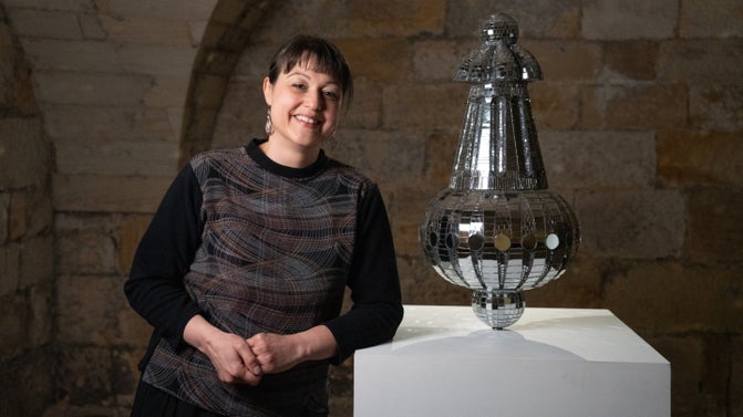 A woman in dark clothing stands leaning on a white exhibition plinth. On the plinth is a salt shaker shape covered in mirrored mosaic tiles.